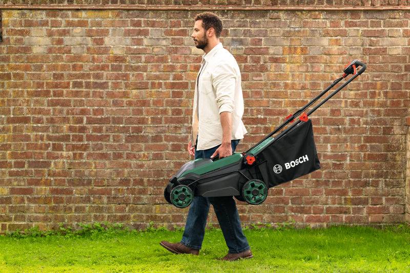 Ein Mann in legerer Kleidung schiebt einen grün-schwarzen Bosch-Rasenmäher über eine grasbewachsene Fläche, mit einer Backsteinmauer im Hintergrund.