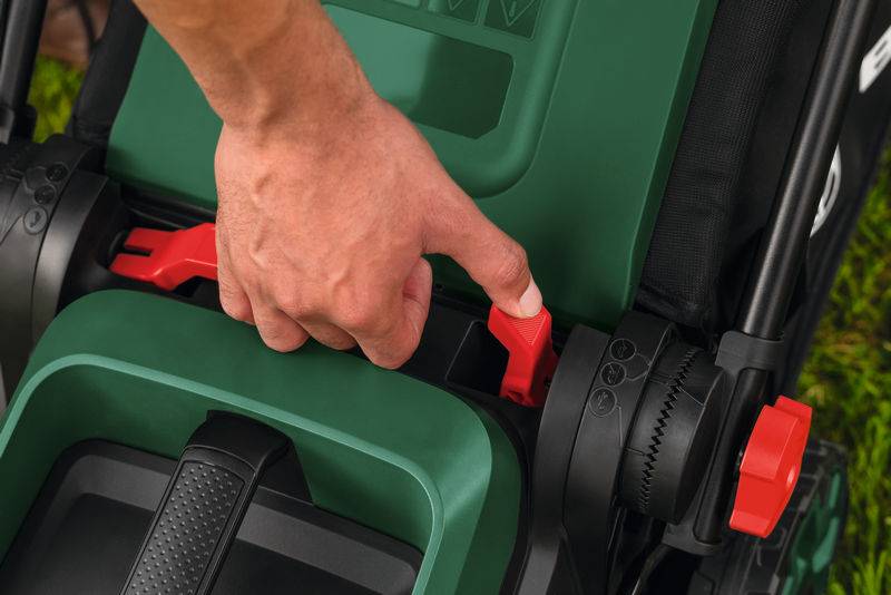 „Eine Hand verstellt die Höheneinstellung eines grünen Rasenmähers mit einem roten Hebel, was auf Benutzerfreundlichkeit und ergonomische Designmerkmale hindeutet."