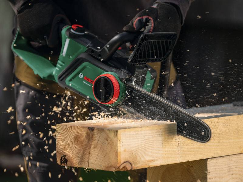 Eine Person nutzt eine Bosch-Elektrokettenäge, um durch ein Holzbrett zu sägen, wobei Holzspäne umherfliegen und den effektiven Werkzeugeinsatz veranschaulichen.