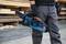 Eine Person hält eine blau-schwarze Kettensäge. Sie trägt graue Hosen und Handschuhe. Holzbretter und Baumstämme sind im Hintergrund sichtbar.