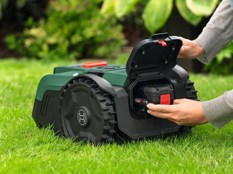 Eine Person setzt eine Batterie in einen Roboterrasenmäher auf einem grünen Rasen ein. Das offene Fach zeigt, wie eine Batterie mit rotem Griff eingesetzt wird.