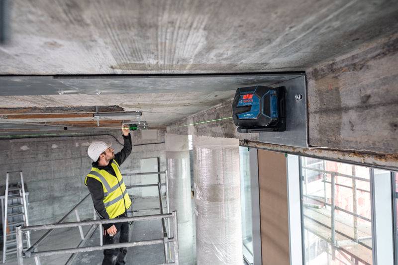 Ein Bauarbeiter in einer Sicherheitsweste und einem Schutzhelm verwendet ein Lasernivelliergerät an einer Betondecke, umgeben von Gerüsten in einem Gebäude.