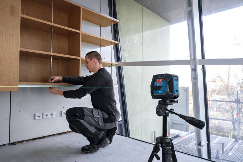 Eine Person, die ein Holzregal mit einem Laser-Messwerkzeug in einem modernen, mit Glaswänden versehenen Raum misst.