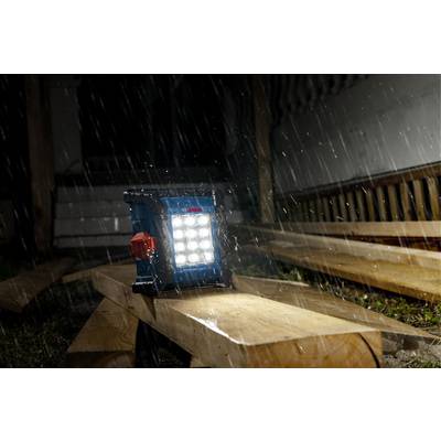 Ein tragbares LED-Arbeitslicht beleuchtet eine Baustelle nachts bei starkem Regen und hebt Holzbretter und teilweise errichtete Strukturen hervor.