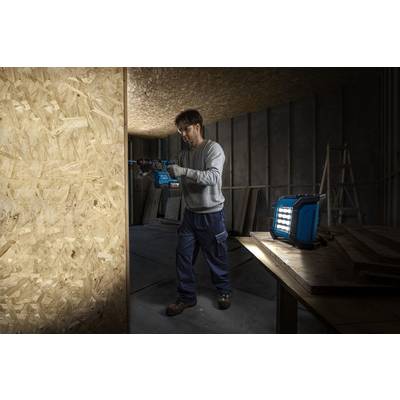 Eine Person in einem schwach beleuchteten Baubereich, die eine Elektrobohrmaschine auf einer Holzplatte verwendet. Eine helle Arbeitsleuchte beleuchtet den Arbeitsbereich.