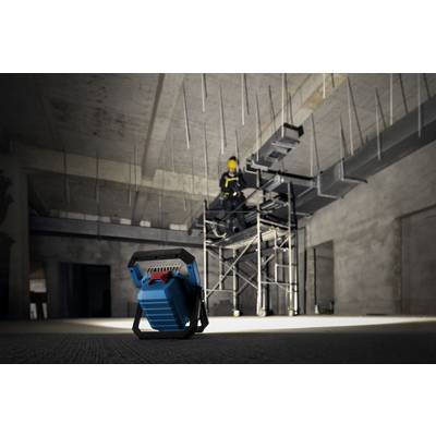 Eine Person auf Gerüsten in einem großen, schwach beleuchteten Industrieraum, mit Fokus auf ein tragbares blaues Arbeitslicht im Vordergrund.