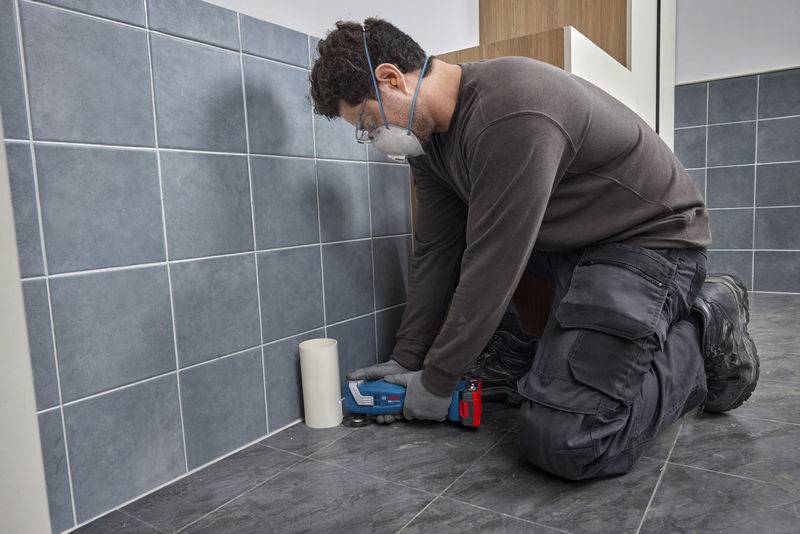 Eine Person kniet auf einem gefliesten Boden, trägt Schutzausrüstung und trägt mit einer Fugenpistole ein Dichtemittel an der Wand an der Basis auf.