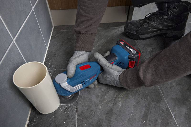 Eine Person nutzt ein blaues Elektrowerkzeug zum Schneiden von Bodenfliesen in der Nähe eines Rohrs und trägt zum Schutz Handschuhe und Stiefel.