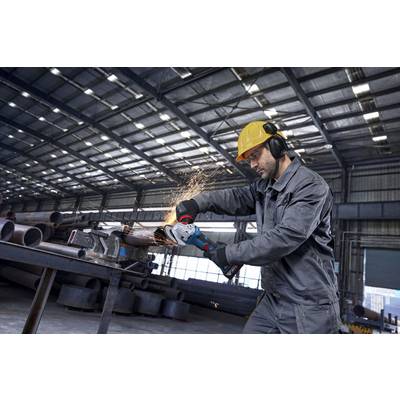 Ein Arbeiter in Schutzausrüstung verwendet einen Winkelschleifer an einem Metallrohr in einem Industrielager und erzeugt dabei Funken.