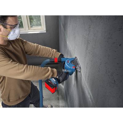 Eine Person trägt Schutzausrüstung, einschließlich einer Maske und Schutzbrille, und verwendet ein Elektrowerkzeug, um eine gerade Linie auf einer Betonwand in einem Innenraum zu schneiden.