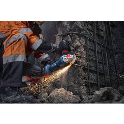 Ein Bauarbeiter in orange Schutzausrüstung benutzt einen Winkelschleifer, um Stahlbewehrung zu schneiden, die aus einer rauen Betonwand herausragt, wobei Funken fliegen.