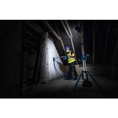Ein Bauarbeiter in einer leuchtend farbigen Warnweste und mit Schutzhelm nutzt ein Elektrowerkzeug, um in einem dunkel beleuchteten Dachboden in eine Wand zu bohren.