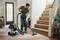 Eine Person saugt einen Holzboden neben einer Treppe in einem hell beleuchteten Wohnzimmer mit einem grün-schwarzen Staubsauger ab.