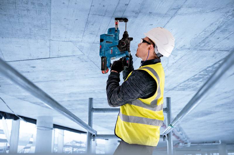 Ein Bauarbeiter in einer gelben Weste und einem Schutzhelm benutzt eine Elektrobohrmaschine an einer Betondecke, was auf aktive Bauarbeiten hinweist.