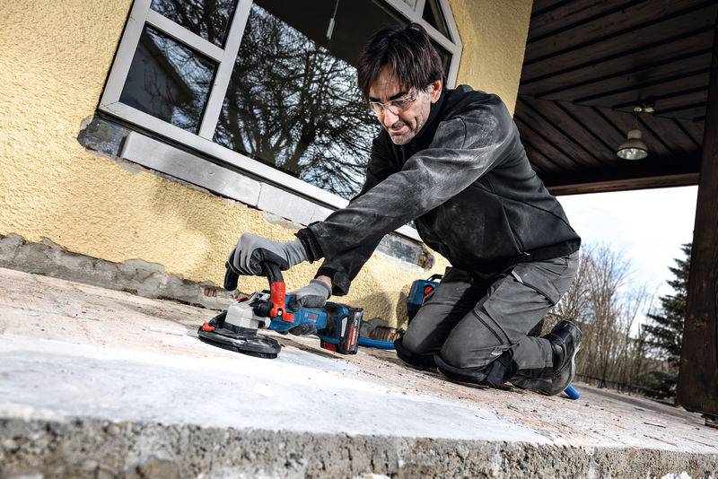Eine Person kniet auf einer Betonveranda und nutzt ein industrielles Elektrowerkzeug, um die Oberfläche zu schleifen. Sie trägt Schutzbrille und Handschuhe.