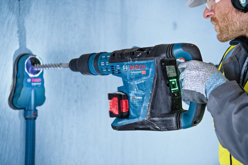 Eine Person benutzt einen Bosch-Schlagbohrer, um ein Loch in eine Wand zu bohren. Der Fokus liegt auf dem Bohrer, der mit einem Staubabsaugsystem ausgestattet ist.