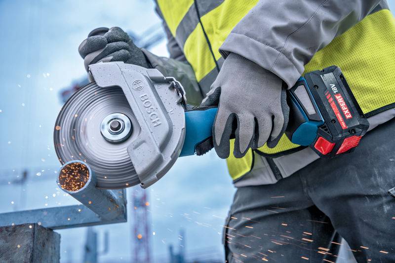 Ein Arbeiter benutzt einen Winkelschleiffer auf Metall und erzeugt dabei Funken. Die Person trägt Schutzhandschuhe und eine neonorange Sicherheitsweste auf einer Baustelle.