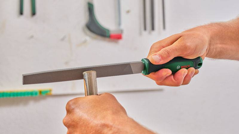 Eine Person nutzt eine flache Handfeile mit grünem Griff, um einen runden Metallstab in einer Werkstattumgebung zu glätten.