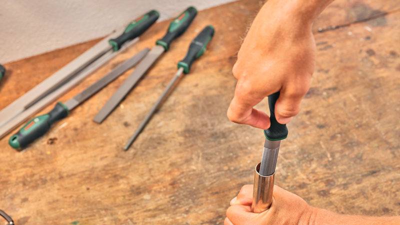 Eine Person dreht einen Schraubendreher in ein zylindrisches Metallobjekt auf einer Holzoberfläche, während verschiedene Werkzeuge in der Nähe liegen.