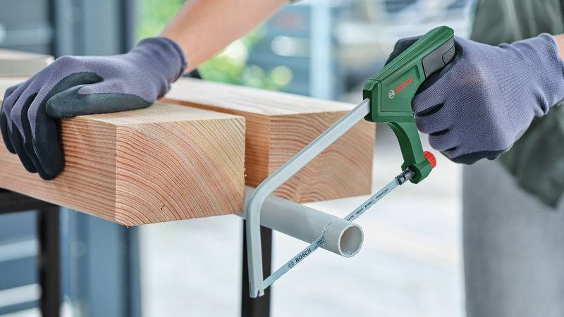 Hände in Handschuhen, die eine grün-schwarze Handsäge verwenden, um ein graues Metallrohr durchzuschneiden, während das Rohr mit der anderen Hand stabilisiert wird.