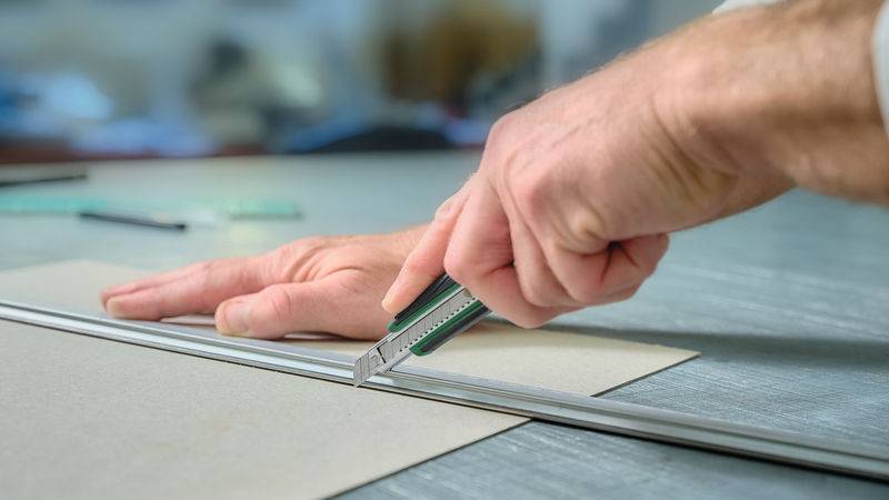Die Hand einer Person benutzt ein Cuttermesser, um ein Stück Pappe mit Hilfe eines Metalllineals auf einem Tisch zu schneiden, wobei der Fokus auf Präzision und Sicherheit liegt.