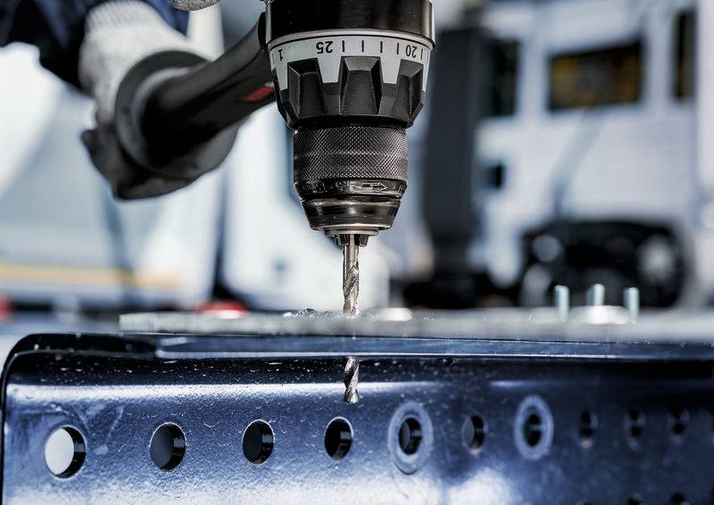 Ein Bohrer dringt in ein Metallblech ein und erzeugt ein Loch. Im Hintergrund ist ein unscharfes Fahrzeug sichtbar, das auf eine Industrieumgebung hindeutet.