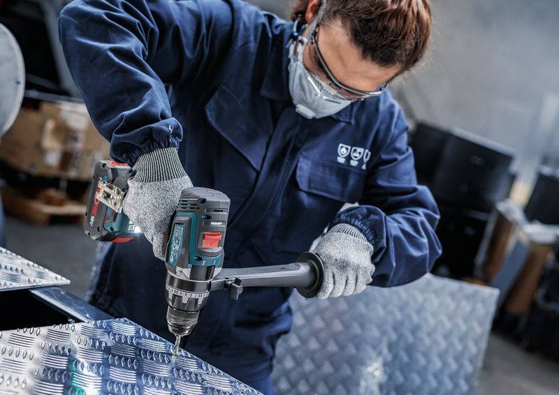 Eine Person in Schutzausrüstung bedient einen elektrischen Bohrer auf einer Metalloberfläche in einer industriellen Umgebung.