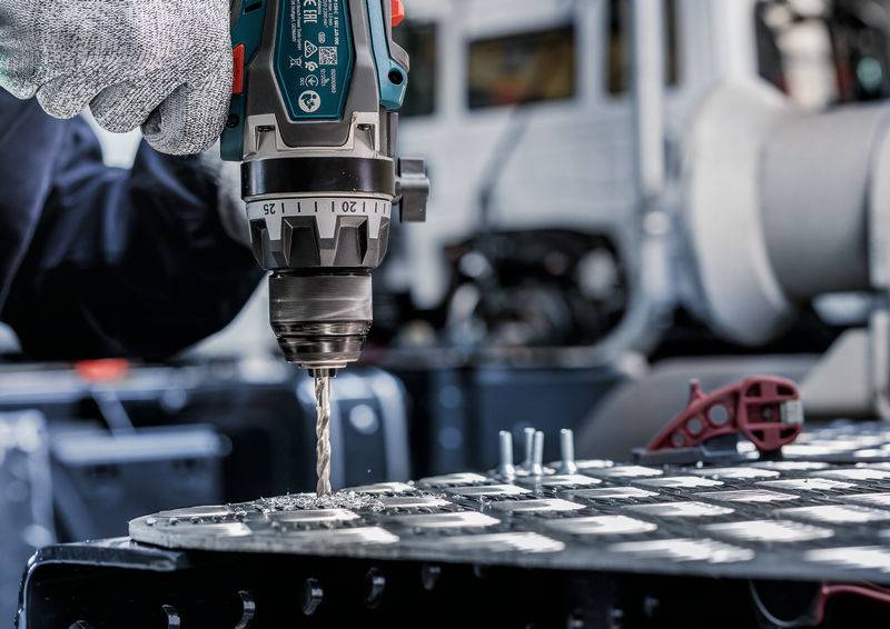 Eine Nahaufnahme einer Person, die eine Elektrobohrmaschine verwendet, um Löcher in Metallplatten zu bohren. Der Bohrer ist fokussiert, und der Hintergrund zeigt eine Werkstattumgebung.