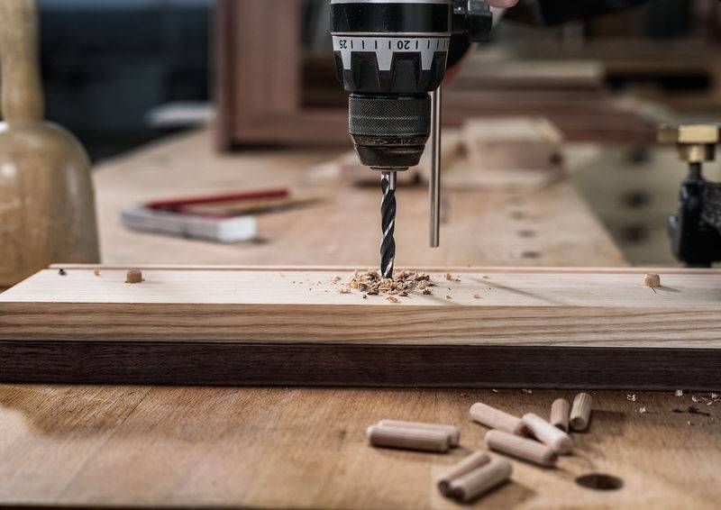 Nahaufnahme einer Bohrmaschine, die in ein Holzbrett auf einer Werkbank eindringt, mit Holzspänen rundherum, Werkzeugen und Dübeln im Hintergrund.