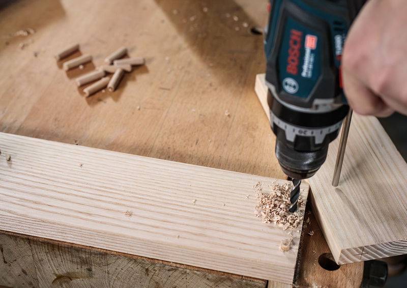 'Person, die ein Loch in ein Holzbrett bohrt, umgeben von Holzdübeln auf einer Werkbank, was auf eine Holzbearbeitungstätigkeit hinweist.'