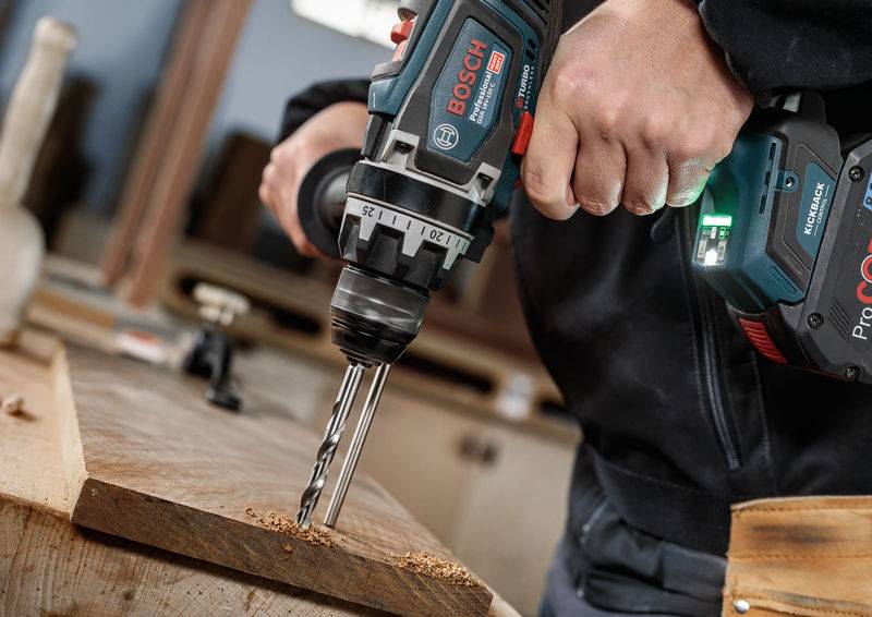 Eine Person, die einen Bosch-Elektroschrauber verwendet, um ein Loch in ein Holzbrett zu bohren und dabei auf einer Werkbank Sägespäne erzeugt.