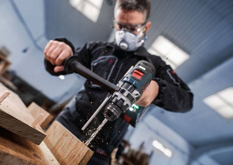 Eine Person, die eine Schutzmaske trägt, bedient einen Elektroschrauber und bohrt in ein Holzbrett in einer Werkstatt, wobei Holzspäne sichtbar sind.