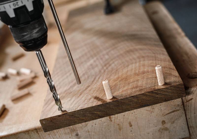 Eine Nahaufnahme eines Bohrers, der Löcher in ein Holzbrett bohrt, mit zwei teilweise eingesetzten Holzdübeln, die die Holzbearbeitung beim Zusammenbau veranschaulichen.