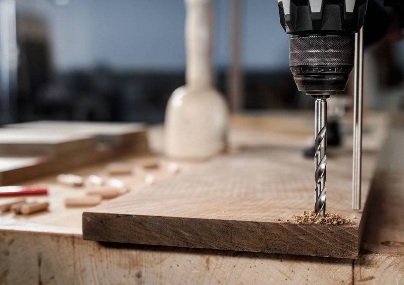 Eine Nahaufnahme einer elektrischen Bohrmaschine, die in ein Holzbrett auf einer Werkbank in einer Werkstättenumgebung mit verstreuten Werkzeugen im Hintergrund bohrt.