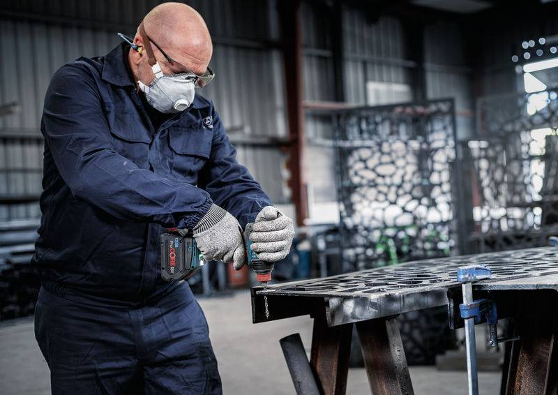 Ein Arbeiter in Schutzausrüstung schneidet Metallbleche mit einem Elektrowerkzeug in einer Industriewerkstatt. Licht fällt durch gemusterte Metallpaneele im Hintergrund.