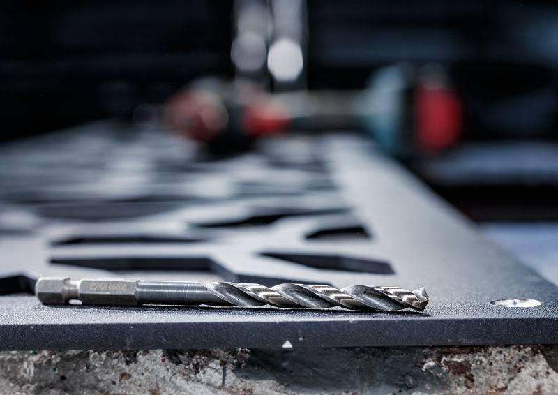 Eine Nahaufnahme eines Bohrers, der auf einer strukturierten Metalloberfläche ruht, mit unscharfen Werkzeugen, die im Hintergrund sichtbar sind und eine Werkstattumgebung suggerieren.
