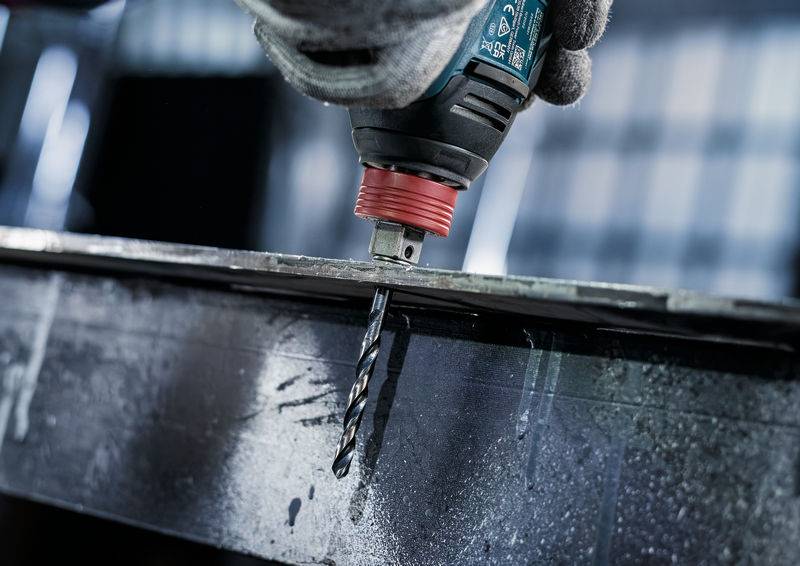 Eine Nahaufnahme einer Hand, die eine Elektrobohrmaschine mit einem Metallbohrer hält, der in einen Stahlträger bohrt. Metallspäne sind um den Bohrer herum sichtbar.