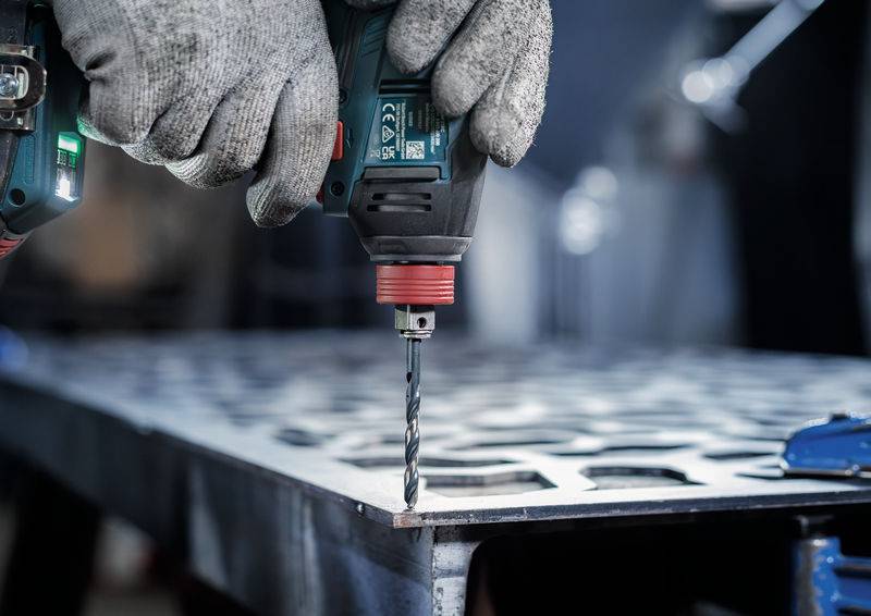 Nahaufnahme von behandschuhten Händen, die eine Elektrobohrmaschine verwenden, um Löcher in ein Metallblech auf einem Werkstatt-Tisch zu bohren.