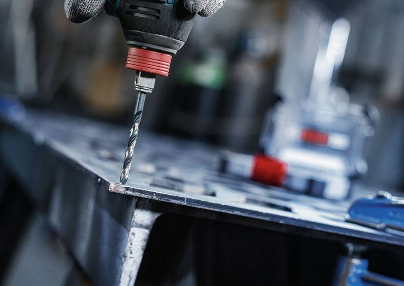 Eine Nahaufnahme einer behandschuhten Hand, die einen kabellosen Bohrer hält und mit Präzision ein Loch in ein Metallblech bohrt; eine Werkstattumgebung im Hintergrund.