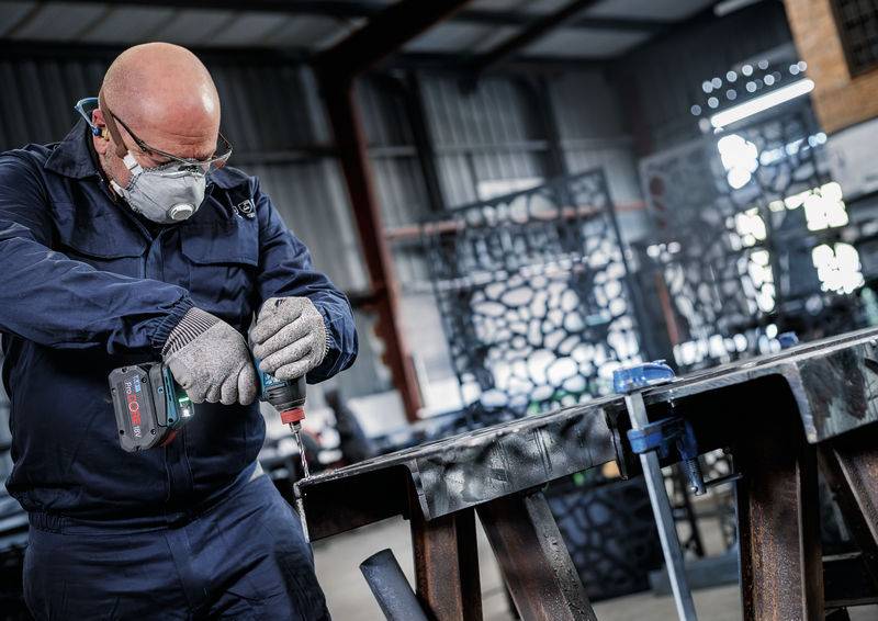 Eine Person in Schutzausrüstung benutzt eine Elektrobohrmaschine an einem Metallträger in einer Industriewerkstatt, mit verschiedenen Werkzeugen und Strukturen im Hintergrund.