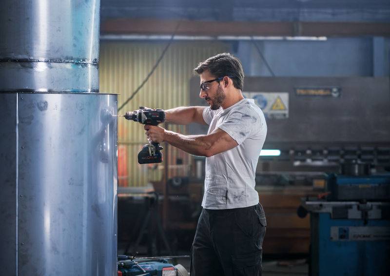 Ein Mann mit Schutzbrille verwendet einen kabellosen Bohrer an einem großen Metallzylinder in einer Industriewerkstatt, umgeben von Maschinen.
