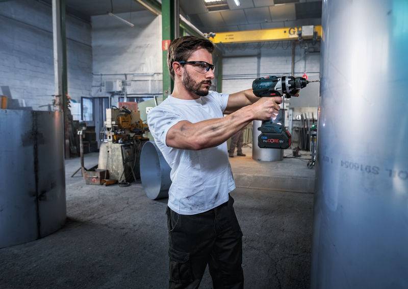 Ein Mann mit Schutzbrille benutzt einen Elektrobohrer an einem großen Metallzylinder in einer Industrieumgebung. Ausrüstung und Maschinen sind im Hintergrund sichtbar.