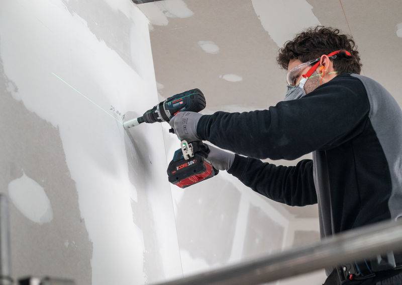Eine Person, die eine Schutzbrille und eine Maske trägt, benutzt einen elektrischen Bohrer, um an Gipskartonplatten in einem Gebäude in Bau zu arbeiten.