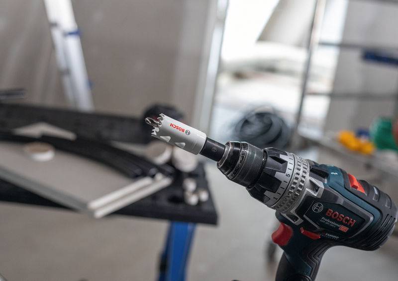 Ein Elektrobohrer der Marke 'Bosch' mit Lochsägen-Aufsatz steht im Fokus, verwendet in einer Baustelle mit verschiedenen Werkzeugen im Hintergrund.