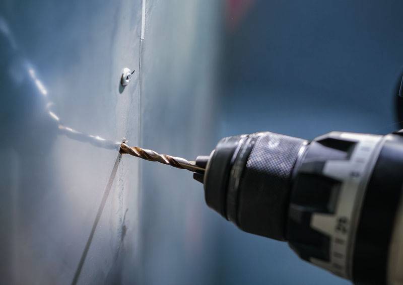 Eine Nahaufnahme eines Elektroschraubers mit einem Metallbohrer, der ein Loch in eine reflektierende Metalloberfläche bohrt und den Bohrprozess hervorhebt.