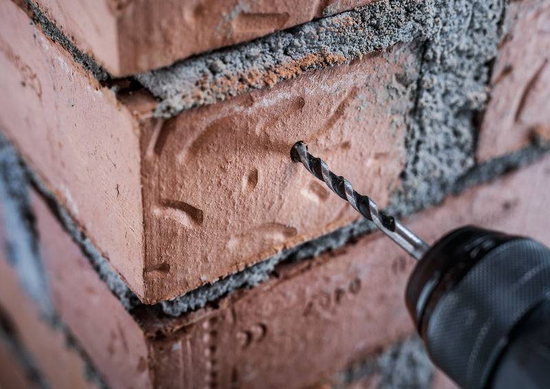 Ein Bohrer macht ein Loch in eine rote Backsteinmauer, Nahaufnahme, zeigt Ziegeltextur und Mörteldetails.