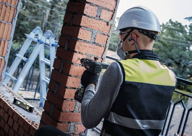 Ein Bauarbeiter in einer Warnweste und einem Helm benutzt einen Bohrer an einer Backsteingestütze; eine Leiter ist im Hintergrund.