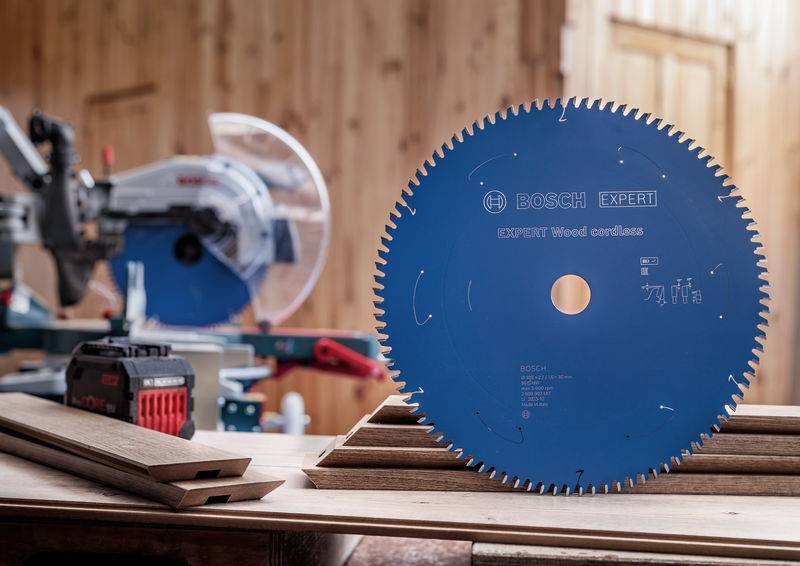 Ein blaues Bosch-Kreissägeblatt mit der Bezeichnung „EXPERT Wood" liegt auf einer Werkbank. Ein Elektrowerkzeug, Holzbretter und ein Akku sind im Hintergrund zu sehen.