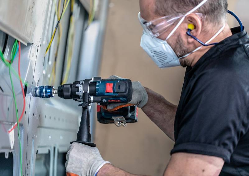 Eine Person, die Schutzausrüstung trägt, einschließlich einer Maske und Schutzbrille, verwendet einen Elektroschrauber an einer Wand mit freiliegenden farbigen Drähten.