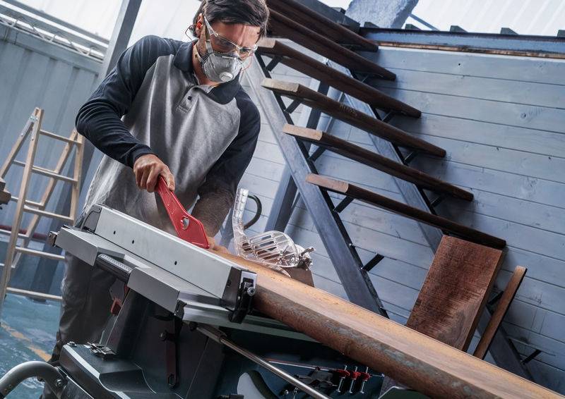 Ein Mann in Schutzausrüstung bedient eine Tischkreissäge in einer Werkstatt und schneidet ein Holzbrett. Im Hintergrund sind Treppen und Werkzeuge sichtbar.